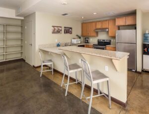 large kitchen island with stools, kitchen with light brown cabinets, stainless steel appliances, cement flooring, and large shelving along wall